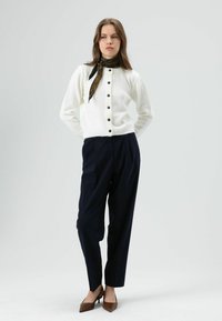 Witte gebreide cardigan met zwarte knopen, gecombineerd met marineblauwe broek. Een donker patroonige sjaal accentueert de halslijn. Bruine puntige hakken maken de outfit compleet.