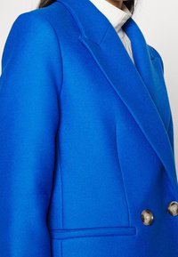 Blazer in lana blu con revers a punta, due bottoni, tessuto testurizzato e tasche frontali. Presenta una vestibilità su misura e spalle strutturate.