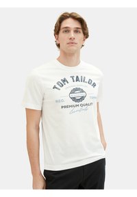 Balts kokvilnas T-krekls ar zilu logo druku, kas uzrāda "TOM TAILOR" un "PREMIUM QUALITY" krūšu daļā. Klasiska apaļā kakla dizains.