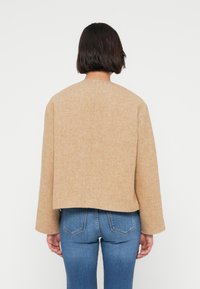 Μπεζ υφασμάτινο σακάκι με ευθεία, κοντή κοπή και χαμηλωμένους ώμους, εμφανιζόμενο από πίσω, σε συνδυασμό με εφαρμοστά μπλε τζιν.