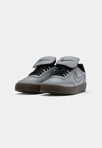 Et par grå Nike-sneakers med sorte snører, sorte Nike-logoer på tungen, og brune gummisåler på en hvit bakgrunn.