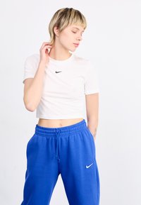 Weißes, kurzes T-Shirt mit schwarzem Nike-Logo, kombiniert mit blauen Jogginghosen, die ein weißes Nike-Logo aufweisen. Schlichter Hintergrund, weicher Stofftextur.
