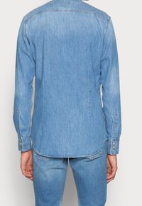 Denimskjorta i ljusblått med långa ärmar och knäppta manschetter. Har en rundad nederkant och en ren design på baksidan.