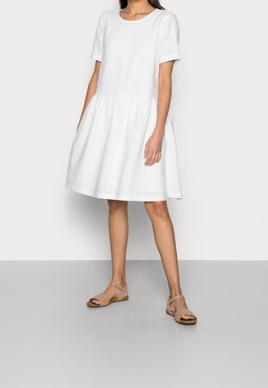 Kvinde iført en løs, kortærmet hvid kjole med samlet talje og beige sandaler med åben tå, stående mod en ensfarvet baggrund.