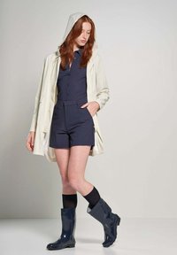 Lichtbeige waterdichte jas met een capuchon, gedragen over een donkerblauwe knoopsluiting top en marineblauwe shorts. Marineblauwe rubberen regenlaarzen maken de look compleet.