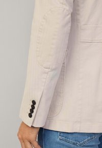 Helle beige Jacke mit tailliertem Schnitt, runden Ellbogenpatches und vier schwarzen Knöpfen an den Bündchen, mit kontrastierenden Steppakzenten.