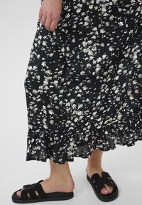 Rochie neagră cu flori și modele albe, având un tiv volănaș. Poartă încălțări negre cu talpă plată și curea lată.