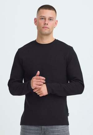 T-shirt noir à manches longues et col rond en tissu texturé, avec un logo subtil sur la poitrine, associé à un jean gris.