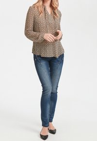 Brun mönstrad blus med geometriska mönster, kombinerad med mörkblå smala jeans. Blusen har en volang i halsringningen, åtsittande ärmar och knappdetaljer.