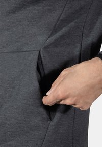 Grå hettegenserstoff med en sidelomme, som viser en hånd som strekker seg inn. Har en myk tekstur og skrå sømdesign.