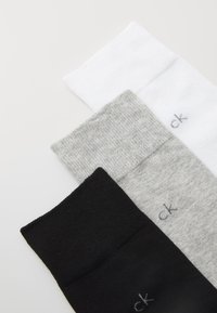 Drei Paar Socken: schwarz, grau und weiß. Jede hat einen gerippten Bund und eine gestrickte Textur mit einem kleinen in grau gestickten Logo.