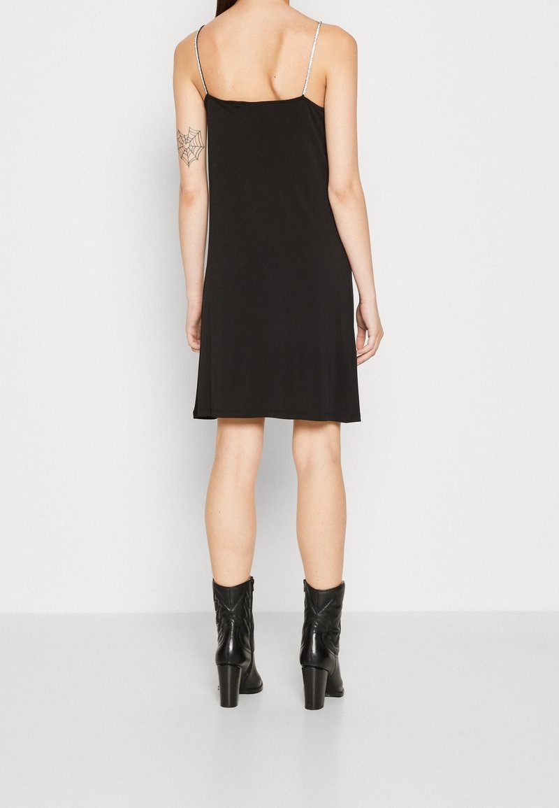 Femme portant une robe noire sans manches avec de fines bretelles et des bottines noires à talons, montrant son dos et un tatouage de toile d'araignée sur son bras.