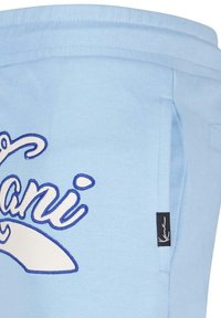 Pantalones de chándal azul claro con una textura suave, que presentan letras bordadas en blanco y azul y un bolsillo lateral. Etiqueta de la marca negra en el lateral.