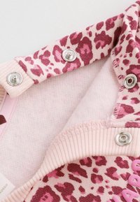 Rosa och mörkröd leopardmönstrad tyg med metallknappar på ribbade rosa kanter, visar en närbild av plaggets beslag.