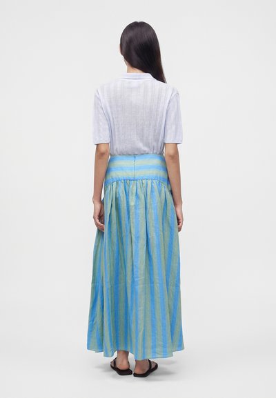Femme debout, de dos, portant un haut blanc à manches courtes et une longue jupe rayée bleue et verte avec des sandales noires.