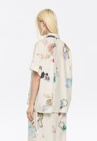 Camicetta a maniche corte di colore crema con stampe botaniche e animali multicolori. Presenta una vestibilità comoda e colletto, realizzata in tessuto di cotone leggero.
