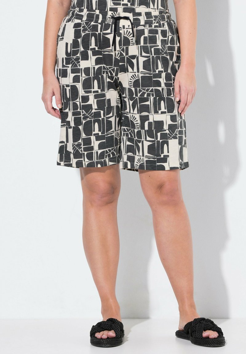 Pantalones cortos de patrón en blanco y negro con cinturilla elástica y cordón, presentan un diseño geométrico y un corte hasta la rodilla. Combinados con sandalias negras.