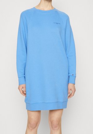 Kobieta nosząca długą, sięgającą kolan niebieską sukienkę z długim rękawem oraz logo Tommy Hilfiger na piersi i mankiecie.