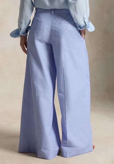 Άτομο που φοράει ανοιχτό μπλε παντελόνι με φαρδιά πόδια και ανοιχτό μπλε πουκάμισο με μακριά μανίκια, στέκεται με τα χέρια στις τσέπες μπροστά σε απλό φόντο.