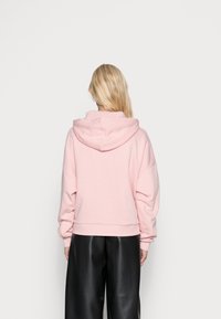 Růžová mikina s kapucí vyrobená z měkké látky, s uvolněným střihem a spadenými rameny, kombinovaná s černými volnými kalhotami.