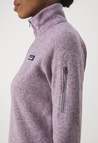 Polaire violette à col montant, avec une poche zippée sur la manche gauche et un petit logo sur la poitrine. Texture lisse.