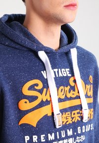 Sudadera azul marino con cordones blancos, que presenta un texto en naranja y blanco "Super Dry" y "Vintage" en una fuente bold y vintage.