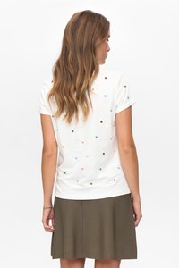 Eine Frau mit langen braunen Haaren trägt ein weißes Kurzarmshirt mit mehrfarbigen Punkten und einen olivgrünen Rock und ist von hinten zu sehen.