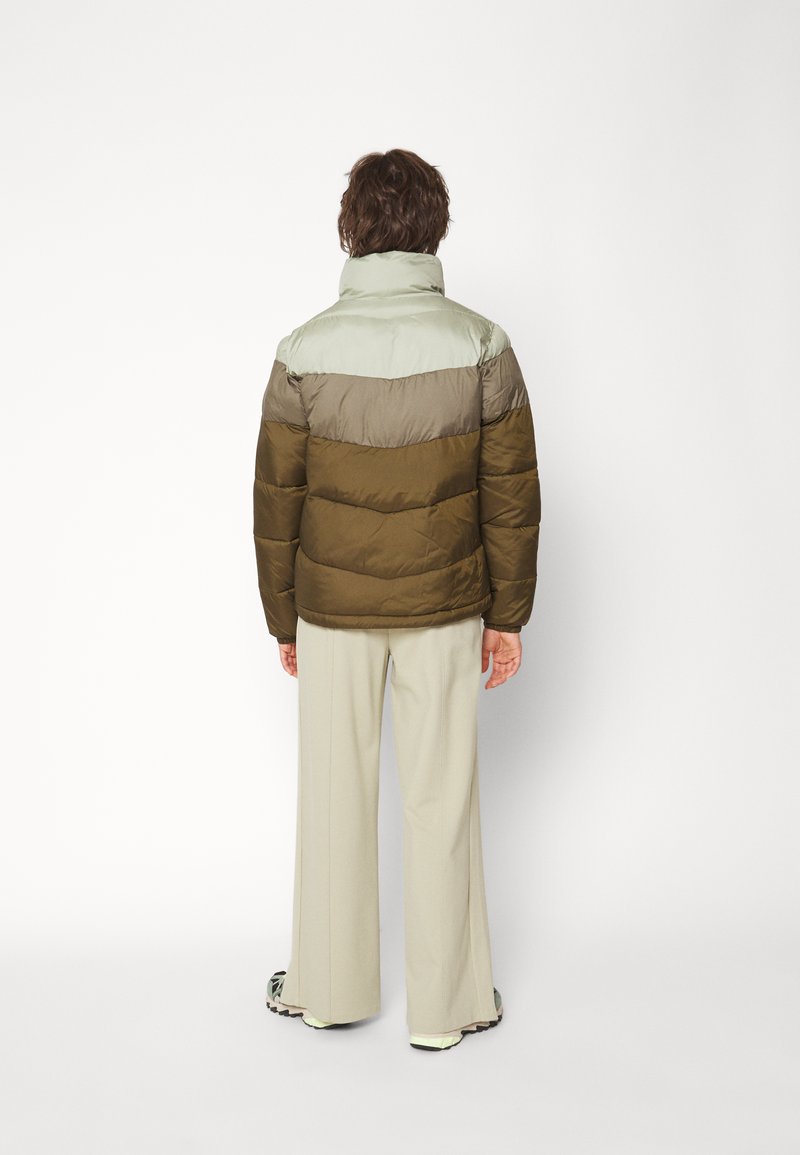 Chaqueta tipo puff con la parte superior de un verde claro y la parte inferior de un verde oliva. Presenta un cuello alto y una textura acolchada.