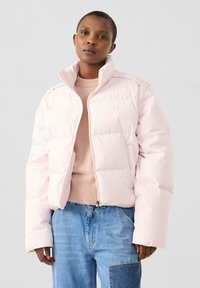 Lichtroze donsjack met een hoge kraag, voorzien van een ritssluiting en een gewatteerd ontwerp. Gedragen over een beige trui en blauwe spijkerbroek.