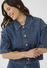 Hymyilevä nainen, jolla on pitkät ruskeat hiukset, pukeutuneena lyhythihaiseen, napilla varustettuun siniseen farkkutakkiin, kädet ristissä tylsää taustaa vasten.