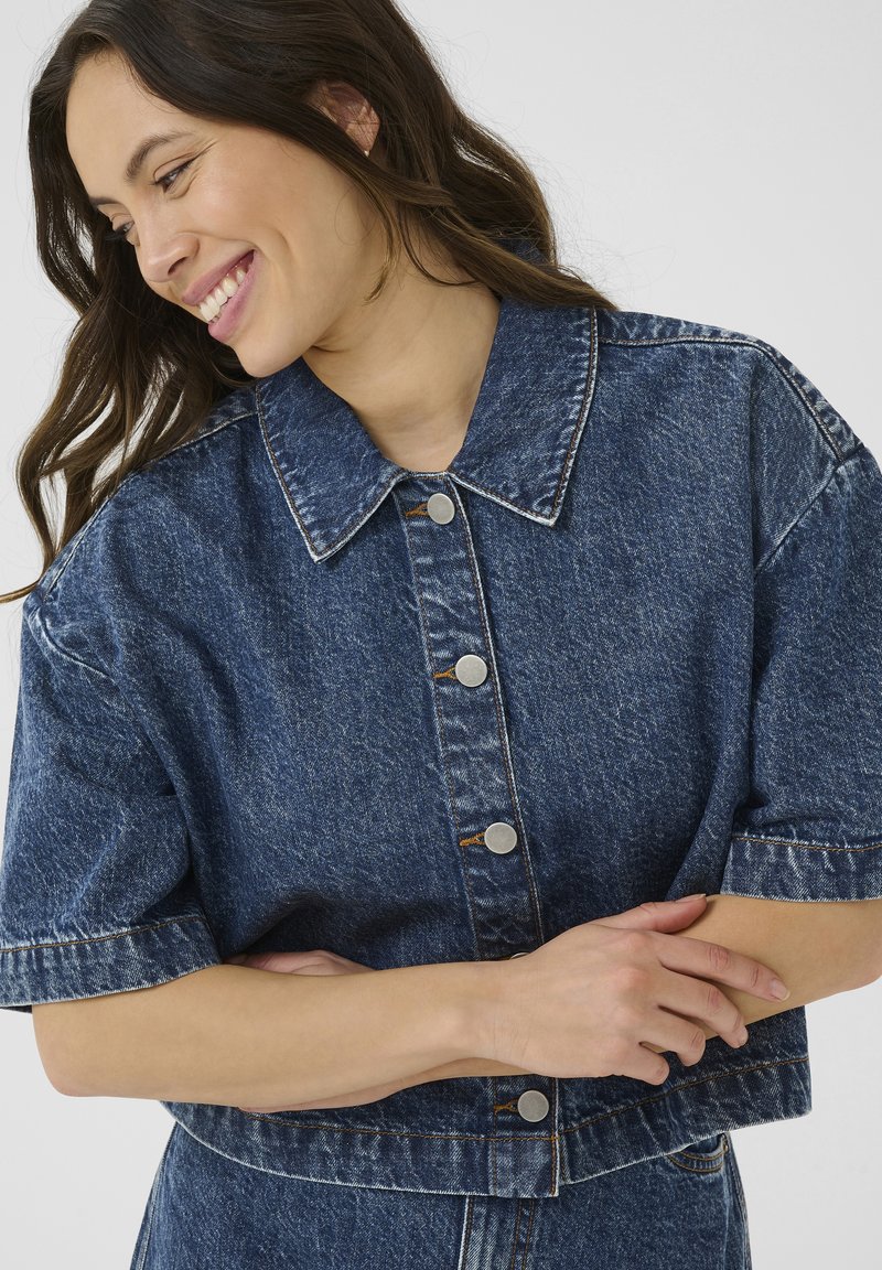 Hymyilevä nainen, jolla on pitkät ruskeat hiukset, pukeutuneena lyhythihaiseen, napilla varustettuun siniseen farkkutakkiin, kädet ristissä tylsää taustaa vasten.