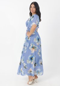 Maxikleid aus hellblauem Stoff mit großen blumenartigen Mustern in Weiß und Dunkelblau. Elastischer Bund, kurze Ärmel, luftiges Design, kombiniert mit weißen Absätzen.
