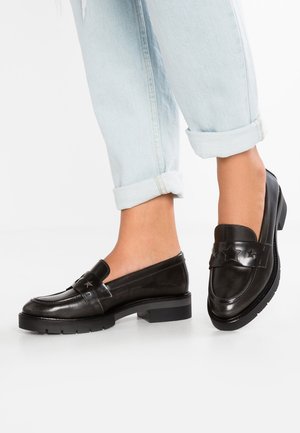 Sorte læder loafers med en blank finish, der har en dekorativ rem med stjerne detaljer og en lav, chunky hæl. Båret med lyseblå, opkrøllede jeans.