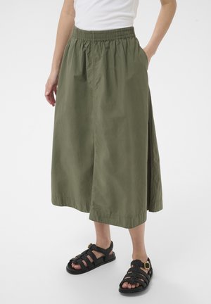 Donna che indossa una gonna lunga fino alla caviglia di colore verde oliva con cintura elastica, top bianco e sandali neri intrecciati con fibbie.