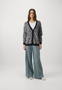Cardigan con motivo geometrico in nero e grigio, dotato di scollatura a V profonda, maniche lunghe e chiusura a bottoni. Abbinato a pantaloni in denim a gamba larga.