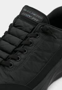 Scarpa Skechers nera con parte superiore trapuntata, dotata di lacci rotondi neri, colletto imbottito e etichetta "Cozy Fit" sulla linguetta.