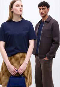 Γυναίκα με μπλε πουκάμισο και κίτρινη καρό φούστα κρατάει μπλε τσάντα χειρός, άντρας με καφέ σακάκι και παντελόνι με μπλε πουκάμισο στέκεται κοντά.