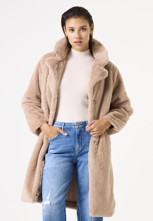 Manteau en fausse fourrure kaki avec un col montant et une fermeture à boutons, porté par-dessus un haut beige et un jean bleu. Texture douce avec une coupe décontractée.