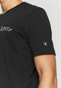 Close-up of a person wearing a black short-sleeve shirt with white text on the chest and a small white logo on the sleeve.