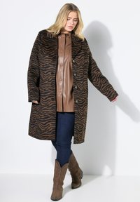 Bruin en zwart zebra-print jas met grote revers en knoopsluiting, gedragen over een bruin faux leren top en donkerblauwe jeans.