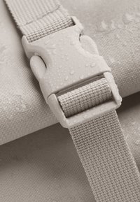 Sangle imperméable beige avec un tissage texturé, dotée d'une boucle en plastique durable, avec des gouttes d'eau visibles à la surface.