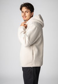 Crème fleece pullover met een capuchon, voorzien van een zachte textuur en zijzakken, gedragen over een donkere jeans. Het model staat iets schuin.