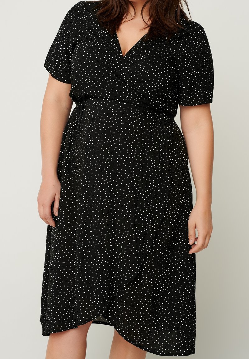 Femme portant une robe portefeuille noire à manches courtes et à pois blancs, debout devant un fond clair uni.