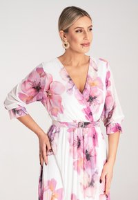 Kwiatowa sukienka na białym tle, zdobiona różowymi i fioletowymi kwiatami. Wykonana w designie z dekoltem w serek, z rękawami trzech czwartej długości oraz detalem w postaci paska w talii.