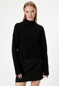 Schwarzer gerippter Rollkragenpullover mit langen Ärmeln, verkürzter Länge und fransigem Saum. Kombiniert mit einem schlichten schwarzen Rock.