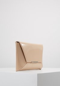 Pochette beige brillante avec un rabat triangulaire et une fermeture rectangulaire métallique, posée sur un socle blanc sur un fond uni.