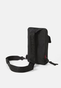 Svart crossbody-veske laget av slitesterkt stoff, med glidelåslukking, justerbar stropp med preget logo og røde detaljer. Rektangulær form.