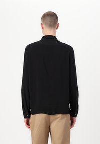 Schwarzes Langarmshirt mit lockerer Passform, mit abgerundetem Saum und geknöpften Manschetten. Glatter Stoff mit einem sanften Glanz, ohne Muster.