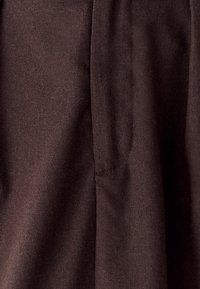 Brauner Stoff mit einer strukturierten Oberfläche, der subtile Nahtdetails und weiche Drapierungen aufweist. Das Material wirkt robust und gleichzeitig leicht.