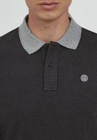Svart polo skjorte med grå krage, med en tre-knapps knapplist og et lite hvitt globus-emblemet på venstre bryst. Strukturert stoff.